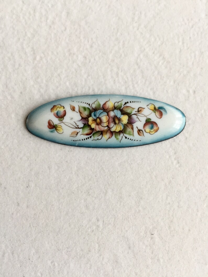 May include: An oval-shaped enamel pin with a blue background and a hand-painted floral design in shades of yellow, pink, and green.