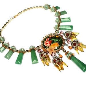 May include: A green and gold beaded necklace with a floral pendant. The pendant features a round, green and gold beaded frame with a floral design inside. The necklace is accented with green and gold beads and a long, green and gold beaded tassel.