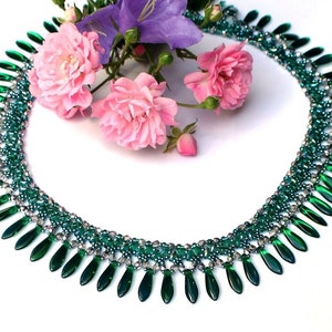 Könnte beinhalten: Eine grüne Perlenkette mit einem kaskadierenden Design aus smaragdgrünen Tropfenperlen. Die Kette liegt auf einem weißen Hintergrund mit rosa Blumen.
