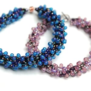 May include: Two beaded bracelets, one in shades of blue and purple, the other in shades of pink and purple.