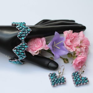 Könnte beinhalten: Ein blaues Perlenarmband in Türkis mit Zickzackmuster und passenden herzförmigen Ohrringen. Der Schmuck wird auf einer schwarzen Hand mit rosa und violetten Blumen präsentiert.
