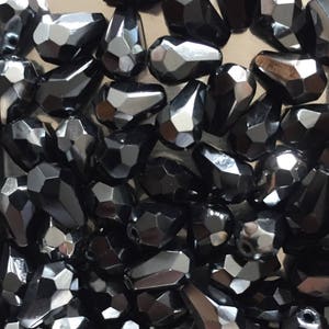 Könnte beinhalten: Nahaufnahme eines Stapels schwarzer, facettierter, tropfenförmiger Perlen. Die Perlen sind aus Glas und haben eine glänzende, reflektierende Oberfläche.