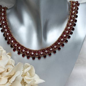 Könnte beinhalten: Eine burgunderrote und goldene Perlenkette mit klaren Kristall-Akzenten. Die Kette befindet sich auf einer Mannequin-Büste.