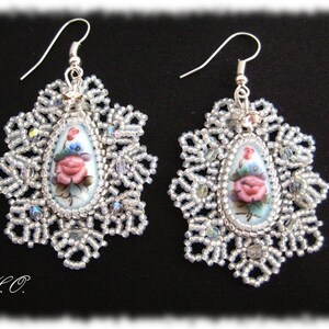 May include: A pair of decorative earrings with a floral motif. Each earring has a central oval pendant with a painted pink rose and blue details, set within a beaded, snowflake-shaped frame. The earrings have silver-coloured hooks.