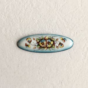 May include: An oval enamel brooch featuring a blue background and a hand-painted floral design in shades of yellow, pink, and green.