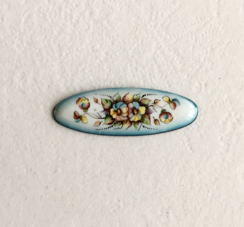 May include: An oval enamel brooch featuring a blue background and a hand-painted floral design in shades of yellow, pink, and green.