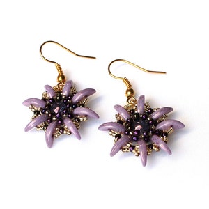 Puede incluir: Un par de pendientes de estrella de color lavanda con ganchos dorados. Los pendientes están hechos con pequeñas cuentas moradas y una cuenta morada más grande en el centro.