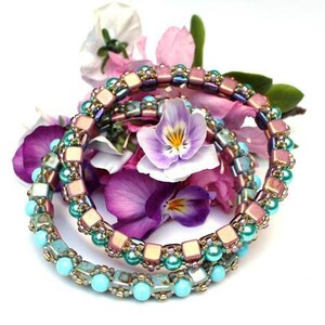 May include: Three beaded bracelets in shades of turquoise, gold, and rose gold. The bracelets are stacked on top of each other and are surrounded by purple and pink flowers.
