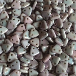 May include: A close-up view of a pile of small, triangular, iridescent glass beads. The beads are a light purple color with a hint of blue.