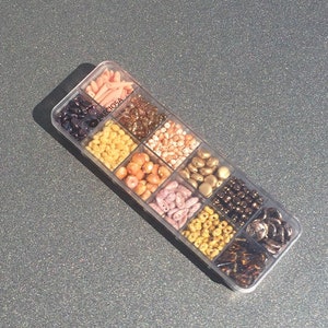 Puede incluir: Una caja de almacenamiento de plástico transparente llena de cuentas de varios colores. Las cuentas son de diferentes formas y tamaños. La caja está etiquetada "PRECiosa" con el texto "The Original Czech Beads".