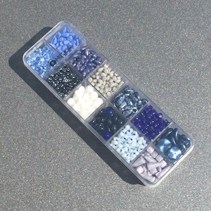 Puede incluir: Una caja de almacenamiento de plástico transparente llena de cuentas de vidrio de varios colores. Las cuentas son de diferentes formas y tamaños. La caja está etiquetada como "Preciosa Czech Beads".