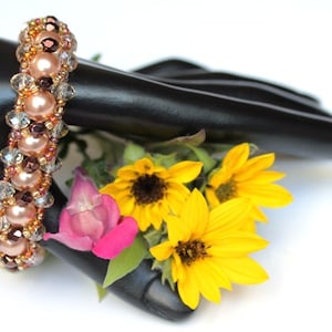 Könnte beinhalten: Ein Armband mit rosa, braunen und klaren Perlen. Das Armband befindet sich auf einer schwarzen Mannequinhand mit gelben und rosa Blumen.