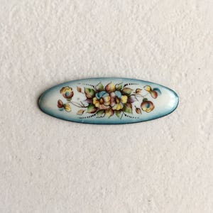 May include: A blue and white oval enamel pin featuring a floral design. The flowers are painted in shades of yellow, pink, and white.