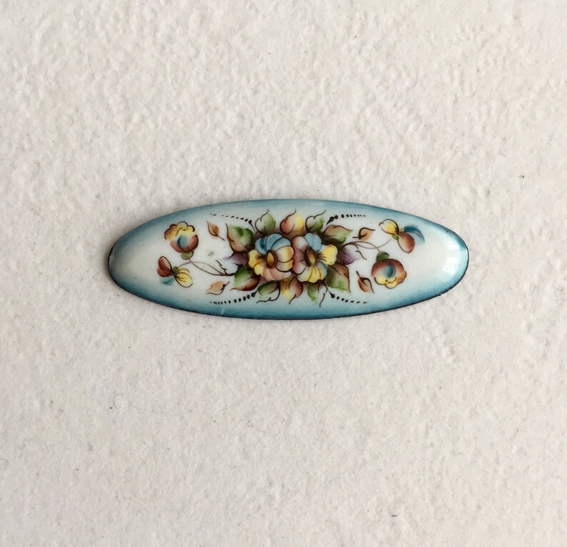 May include: A blue and white oval enamel pin featuring a floral design. The flowers are painted in shades of yellow, pink, and white.