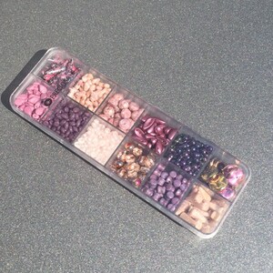 Puede incluir: Una caja de almacenamiento de plástico transparente llena de varios tipos de cuentas en tonos de rosa, morado y marrón. La caja está etiquetada "Preciosa Traditional Czech Beads".