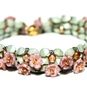 Könnte beinhalten: Ein zartes Armband aus grünen und braunen Perlen mit rosa Blumenakzenten. Das Armband besteht aus mehreren Perlensträngen und hat ein florales Thema.