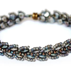 Könnte beinhalten: Ein Armband aus Perlen mit einem mehrfarbigen, irisierenden Finish. Das Armband besteht aus kleinen, runden Perlen in Grautönen, Blau und Braun.