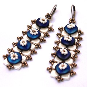 Könnte beinhalten: Ein Paar lange, baumelnde Ohrringe mit einem weißen und blauen Design. Die Ohrringe bestehen aus kleinen Perlen und weisen ein sich wiederholendes Muster aus weißen und blauen Blumen auf.