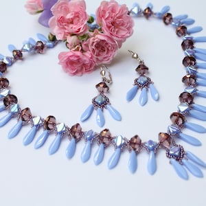 Könnte beinhalten: Eine hellblaue und violette Perlenkette mit passenden Ohrringen. Die Kette ist aus tropfenförmigen Perlen gefertigt und die Ohrringe sind im Hänge-Stil.