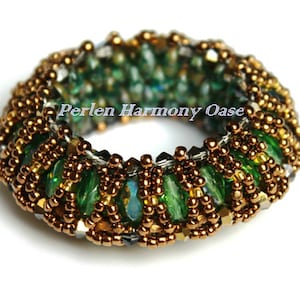 May include: A beaded ring with a detailed design. The ring features a combination of green, gold, and silver beads. The beads are arranged in a circular pattern, creating a textured and eye-catching accessory. The text "Perlen Harmony Oase" is visible.