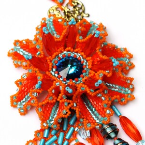 May include: A beaded necklace with a large orange and turquoise flower pendant. The pendant features a large blue crystal in the center. The necklace is made of small beads and has a gold clasp.