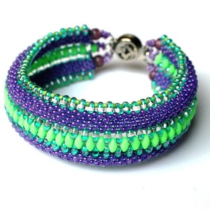 May include: A beaded bracelet with a green, teal, and purple color scheme. The bracelet is made with small seed beads and has a silver clasp.