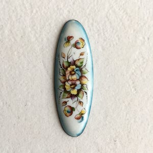 May include: A white oval-shaped enamel pin with a blue border. The pin features a hand-painted floral design with yellow, pink, and purple flowers.