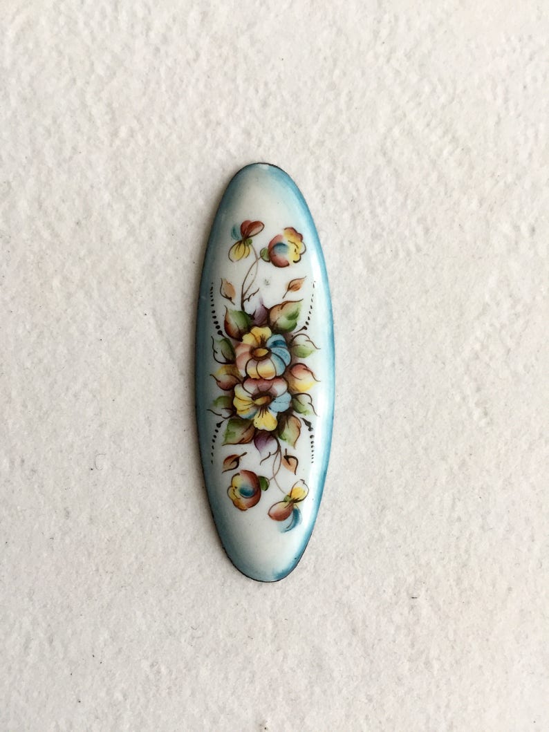 May include: A white oval-shaped enamel pin with a blue border. The pin features a hand-painted floral design with yellow, pink, and purple flowers.