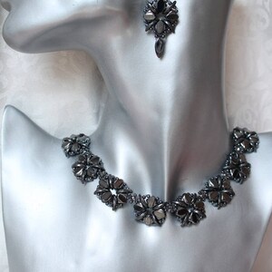 May include: A black necklace and earrings set made of faceted hematite beads. The necklace is a choker style with a row of flower-shaped bead clusters. The earrings are dangle style with a single flower-shaped bead cluster.