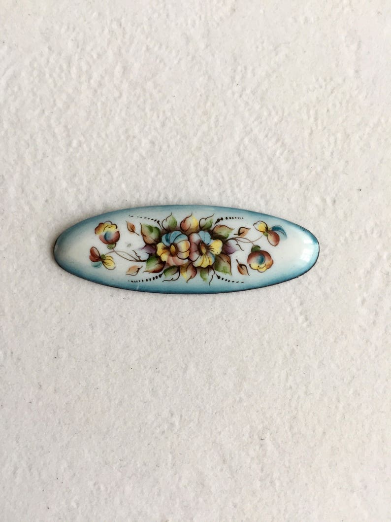 May include: An oval enamel brooch featuring a floral design. The flowers are painted in shades of blue, yellow, and pink, with green leaves and stems. The background is a light blue colour.