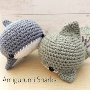 May include: Two crocheted amigurumi shark toys. One is gray with a white belly and the other is a light green color with a white belly. Both have black eyes and are facing the camera. The text "Amigurumi Sharks" is below the toys.
