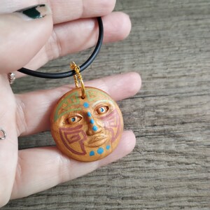 May include: A gold pendant necklace with a round, painted clay pendant featuring a stylized face with blue eyes, red and green lines, and blue dots. The pendant is attached to a black cord.