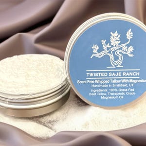 Puede incluir: Dos botes redondos de metal con sebo batido con magnesio. Un bote está abierto, revelando el producto, y el otro está cerrado con una etiqueta azul. La etiqueta dice "Twisted Saje Ranch" y enumera los ingredientes. Los botes están sobre una tela suave de tono neutro.