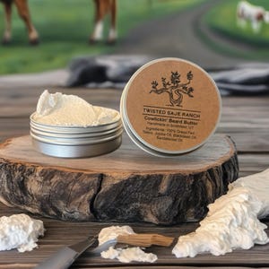 Könnte beinhalten: Nahaufnahme einer silbernen Dose Bartbutter, ohne Deckel, die eine weiße, cremige Substanz zeigt. Das Etikett lautet "TWISTED SAJE RANCH Cowlickin' Beard Butter". Die Dose steht auf einer Holzscheibe, mit einem Messer und mehr Butter in der Nähe. Eine Kuh ist im Hintergrund zu sehen.