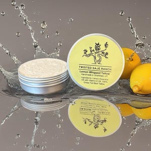 May include: Two open silver tins of Lemon Whipped Tallow with a yellow label and the text "TWISTED SAJE RANCH". The ingredients include Lemon Essential Oil and Grass Fed Beef Tallow. Two lemons are next to the tins.