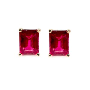 May include: A pair of stud earrings featuring rectangular emerald-cut red gemstones set in gold metal prongs. Each earring displays a vibrant, deep red color with clear facets that reflect light, secured by four small gold corner prongs.