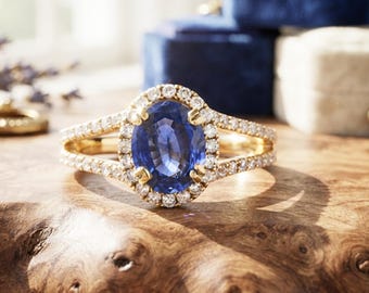 May include: A gold ring featuring a central oval-cut blue sapphire surrounded by a halo of small round white diamonds. The band is split into two delicate arms, each encrusted with a row of tiny diamonds, creating a refined and elegant appearance.