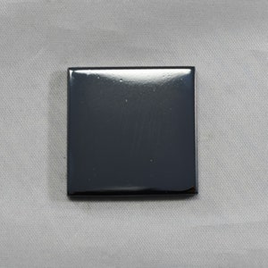May include: A square, dark grey object with a glossy finish. The object is centred and appears to be made of a solid, smooth material. The surface reflects light, creating highlights and shadows. The background is a light grey fabric.