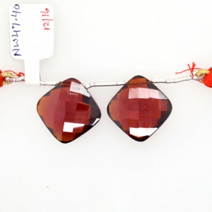 May include: Two red gemstone beads with a faceted square shape. The beads are strung on a silver wire with red tassels on each end. The beads are approximately 12 mm in size.