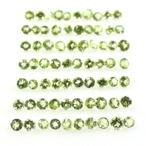 May include: A collection of round, green gemstones arranged in rows on a white surface. The gemstones are all approximately the same size and shape.