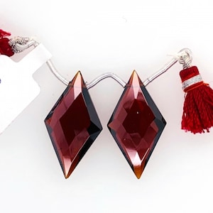 May include: Two faceted, diamond-shaped gemstones in shades of burgundy and amber are strung on a silver wire. A red tassel is attached to one end. The gemstones are displayed against a white background.