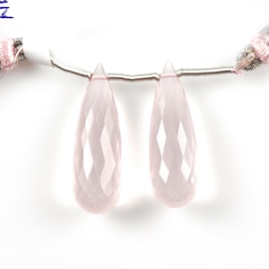 May include: Two faceted, teardrop-shaped rose quartz gemstones. The gemstones are a pale pink color and are strung on a thin, silver-colored wire. The gemstones are displayed against a white background.