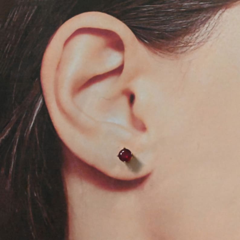 May include: A close-up of a person's ear with a small, round, red gemstone stud earring.