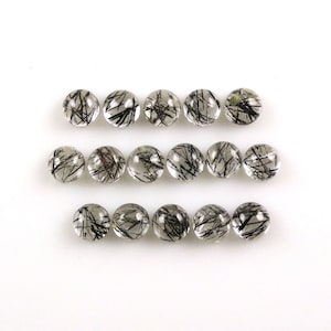 Può includere: Una collezione di 18 cabochon rotondi in quarzo trasparente con inclusioni nere. I cabochon sono disposti in tre file di sei.