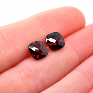 May include: Two red gemstone cushion cut loose stones. The stones are faceted and have a deep red color.