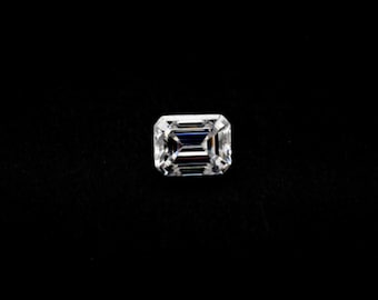 May include: A clear emerald cut diamond gemstone on a black background.