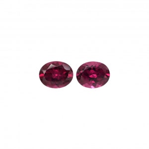 May include: Two oval-cut, deep red gemstones. The gems are set against a white background, showcasing their faceted surfaces and rich color. The stones appear to be of high quality, with a brilliant shine. Ideal for jewelry making or collecting.