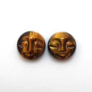 May include: Two round, carved tiger's eye cabochons with a face design. The cabochons are brown and gold with a slight sheen.