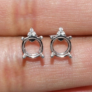 May include: A pair of silver earring settings with a round bezel and three small diamonds on the top of each setting.