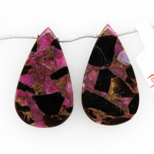Puede incluir: Dos cuentas de piedra preciosa en forma de lágrima con un patrón de mármol negro, rosa y dorado.